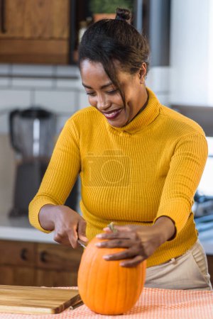 Photo pour Jeune femme noire souriante se préparant pour Halloween sculpter une citrouille à la maison dans la cuisine - image libre de droit