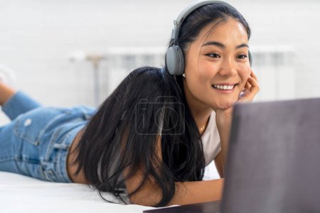Asiatische Studentin, eine junge Frau, die zu Hause die englische Sprache über das Internet lernt. Mit Kopfhörern und Laptop, Teilnahme an einem Online-Kurs