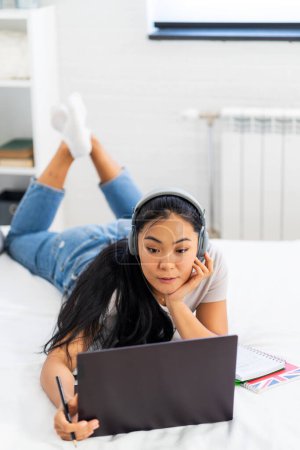 Téléchargez les photos : Étudiante asiatique, une jeune femme apprenant l'anglais sur Internet à la maison. Utilisation d'écouteurs et d'un ordinateur portable, assister à un cours en ligne - en image libre de droit