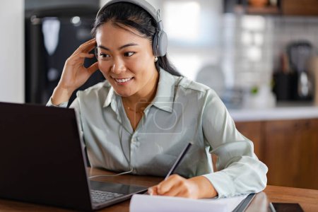 Photo for Young Asian women attending online foreign language classes. Sitting in front of a laptop computer with headphones, listening to courses and taking notes. - Royalty Free Image