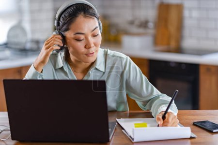 Photo for Young Asian women attending online foreign language classes. Sitting in front of a laptop computer with headphones, listening to courses and taking notes. - Royalty Free Image