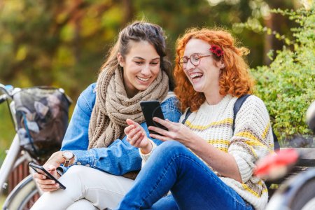 Téléchargez les photos : Deux jeunes femmes assises sur un banc et utilisant un smartphone - en image libre de droit