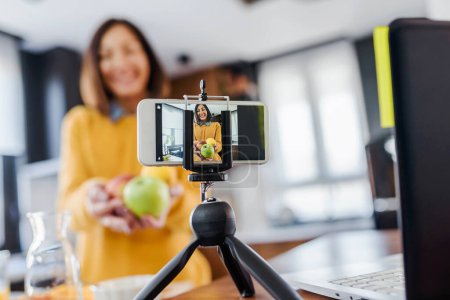 Photo for Young woman making a vlog about healthy food and drinks at her home using mobile phone - Royalty Free Image
