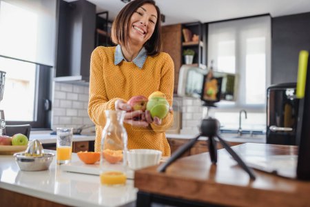 Téléchargez les photos : Jeune femme faisant un vlog sur la nourriture saine et les boissons à sa maison en utilisant un téléphone mobile - en image libre de droit
