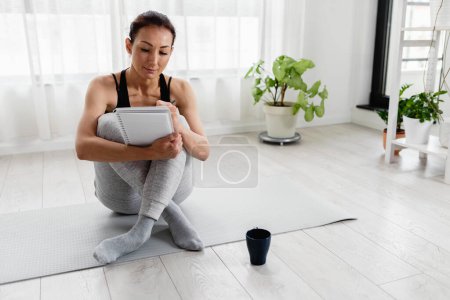 Téléchargez les photos : Jeune femme écrivant dans son carnet. Assis à la maison près de la fenêtre sur un tapis de yoga après l'exercice. Résolutions de croissance personnelle - en image libre de droit