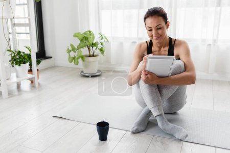Photo for Young woman writing in her notebook. Sitting at home by the window on a yoga mat after exercise. Personal growth resolutions - Royalty Free Image