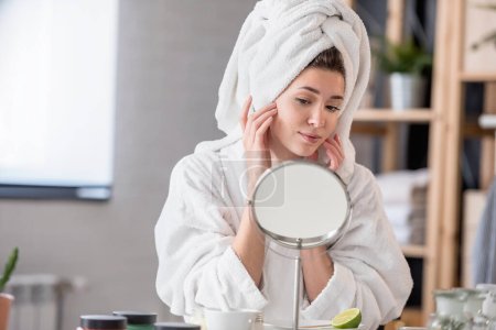 Téléchargez les photos : Belle jeune femme en peignoir et serviette regardant miroir, concept de soins de la peau - en image libre de droit
