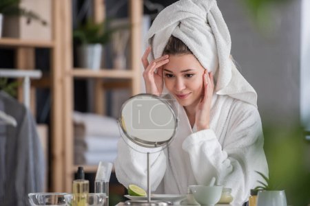 Téléchargez les photos : Belle jeune femme en peignoir et serviette regardant miroir, concept de soins de la peau - en image libre de droit