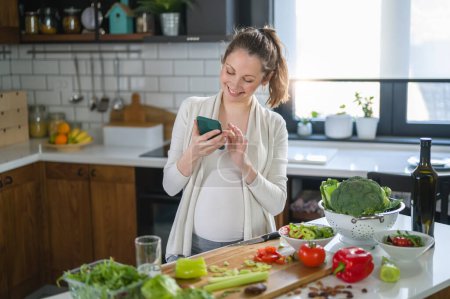 Téléchargez les photos : Belle femme enceinte avec téléphone à la main debout dans la cuisine - en image libre de droit