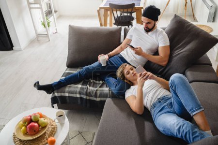 Photo for Couple having rest at home in livingroom, using phones - Royalty Free Image