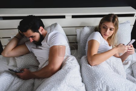 Téléchargez les photos : Jeune couple couché sur le lit et utilisant des téléphones mobiles - en image libre de droit