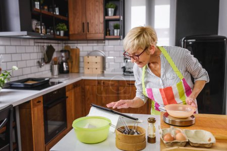 Téléchargez les photos : Femme âgée qui cuisine dans la cuisine de la maison. Utilisation d'un ordinateur tablette pour trouver la recette. Femme mûre cuisine avec l'aide d'Internet. Technologie moderne pour les personnes âgées. - en image libre de droit