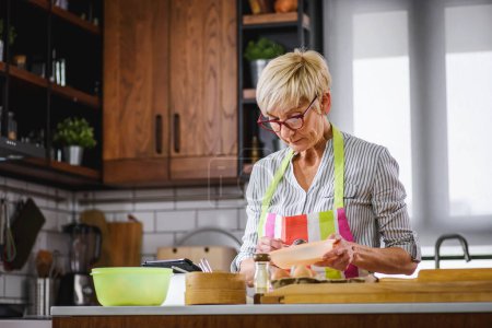 Téléchargez les photos : Femme âgée qui cuisine dans la cuisine de la maison. Utilisation d'un ordinateur tablette pour trouver la recette. Femme mûre cuisine avec l'aide d'Internet. Technologie moderne pour les personnes âgées. - en image libre de droit