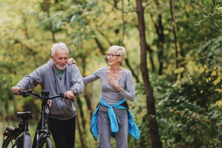 Photo for Cheerful active senior couple with bicycle walking through park together. Perfect activities for elderly people. - Royalty Free Image