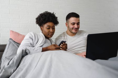 Téléchargez les photos : Jeune couple multiracial utilisant leurs appareils électroniques mobiles au lit. Dépendance à Internet. - en image libre de droit