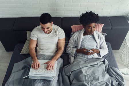 Photo for Young multiracial couple using their mobile electronic devices in bed ignoring each other. Relationship and communication problems and internet social network addiction. - Royalty Free Image