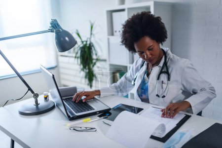 Foto de Doctora que trabaja en su consultorio frente a la computadora portátil, escribiendo un informe de salud - Imagen libre de derechos