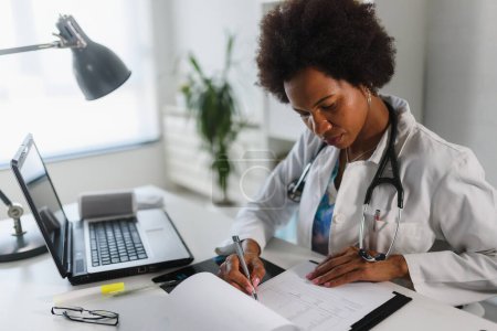 Foto de Doctora que trabaja en su consultorio frente a la computadora portátil, escribiendo un informe de salud - Imagen libre de derechos