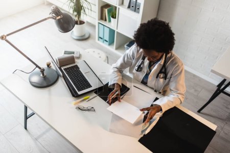 Téléchargez les photos : Femme médecin avec stéthoscope regardant les documents médicaux à son bureau travaillant dur - en image libre de droit