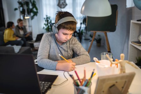 Téléchargez les photos : Concentré garçon sérieux en utilisant un ordinateur portable tout en étant assis sur le bureau à la maison. Technologie, éducation, enseignement à distance concept - en image libre de droit