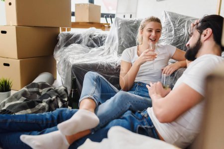 Téléchargez les photos : Joyeux jeune couple emménageant dans une nouvelle maison, assis sur le sol à boire du champagne célébrant - en image libre de droit