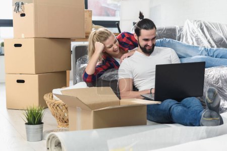 Photo for Young couple moving in new home.Sitting on floor and relaxing after unpacking.Looking something on laptop - Royalty Free Image