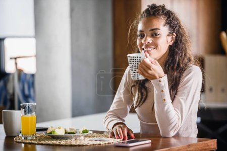 Foto de Joven afroamericana mujer sentada en casa con una taza de café y el uso de teléfono móvil - Imagen libre de derechos