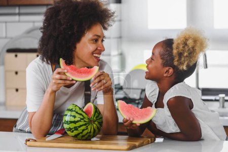 Téléchargez les photos : Heureuse mère afro-américaine et petite fille mangeant pastèque à la maison - en image libre de droit