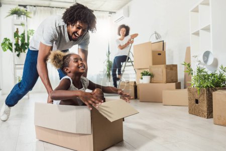 Téléchargez les photos : Heureuse famille afro-américaine ludique déménageant dans un nouvel appartement, petite fille maternelle assise dans des boîtes en carton, père la roulant, concept de propriété d'achat - en image libre de droit