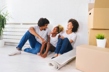 Téléchargez les photos : Jeune famille afro-américaine heureuse déballer pendant le déménagement. Assis sur un plancher reposant à leur nouvelle maison. - en image libre de droit