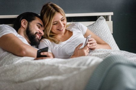 Photo for Happy couple lying in bed and looking at smartphone in morning - Royalty Free Image