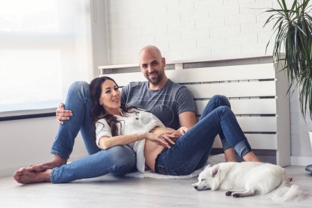 Téléchargez les photos : Jeune couple étreignant dans le salon, chien couché à côté d'eux - en image libre de droit