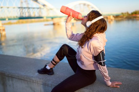 Téléchargez les photos : Jeune femme urbaine se reposant après le jogging, eau potable au bord de la rivière - en image libre de droit