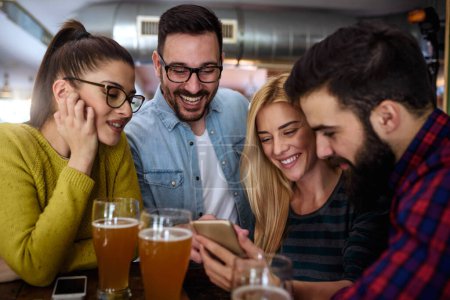 Photo for Friends at the bar drinking beer looking into smartphone - Royalty Free Image