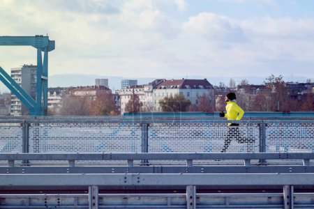 Foto de Hombre atractivo en forma corriendo rápido a lo largo de un gran puente moderno. Ejercicio, trotar, deporte, invierno. Hombre atleta corriendo. - Imagen libre de derechos