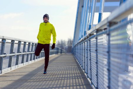 Foto de Joven atleta hombre estirando sus músculos antes de correr en el puente en el día de invierno soleado - Imagen libre de derechos