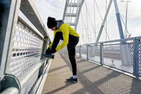 Photo for Young sporty man in windbreaker preparing for jogging over bridge tying his shoes. - Royalty Free Image