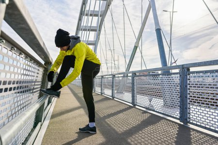 Photo for Young sporty man in windbreaker preparing for jogging over bridge tying his shoes. - Royalty Free Image
