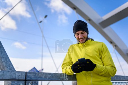 Foto de Retrato de un joven atleta en un rompevientos usando un teléfono inteligente en un día soleado de invierno - Imagen libre de derechos