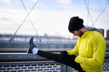 Téléchargez les photos : Jeune athlète homme étirant ses muscles avant de courir sur le pont sur une journée ensoleillée d'hiver - en image libre de droit
