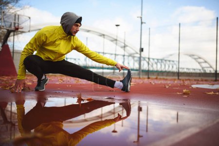 Junger Sportler dehnt seine Muskeln, bevor er an einem regnerischen Tag auf Brücke läuft