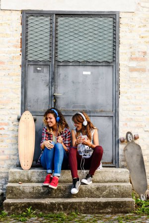 Téléchargez les photos : Skate girls assis dans la rue à écouter de la musique avec écouteurs et smartphone - en image libre de droit