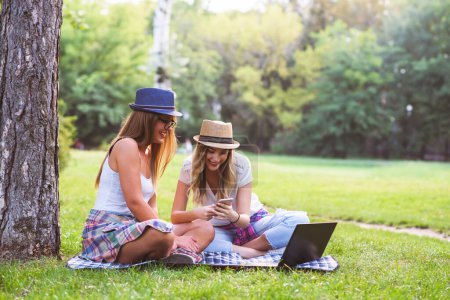 Téléchargez les photos : Deux amies assises sur l'herbe, parlant et utilisant un ordinateur portable - en image libre de droit