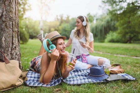 Téléchargez les photos : Filles amis assis dans le parc sur couverture écouter de la musique dans les écouteurs, boire de la limonade - en image libre de droit
