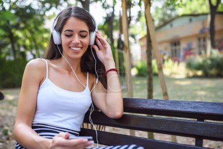 lächelnde Frau hört Musik und sitzt auf Bank im Park