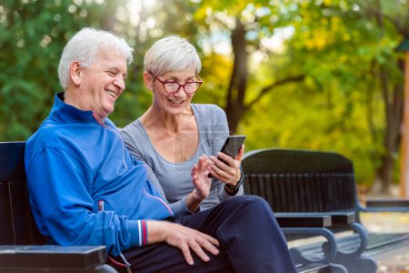 Téléchargez les photos : Couple actif âgé souriant assis sur le banc regardant smartphone - en image libre de droit
