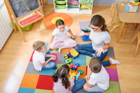 Photo for Preschool teacher talking to group of children sitting on a floor at kindergarten - Royalty Free Image