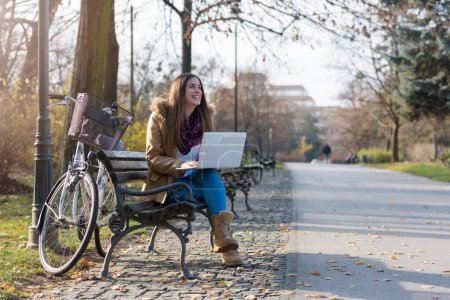 Téléchargez les photos : Étudiante souriante avec vélo sur banc de parc, travaillant sur ordinateur portable - en image libre de droit