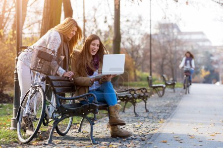 Téléchargez les photos : Groupe de filles souriantes collège sur banc de parc travaillant sur ordinateur portable - en image libre de droit