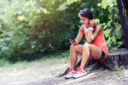 Téléchargez les photos : Sport actif fille assise dans le parc manger pomme écouter de la musique - en image libre de droit